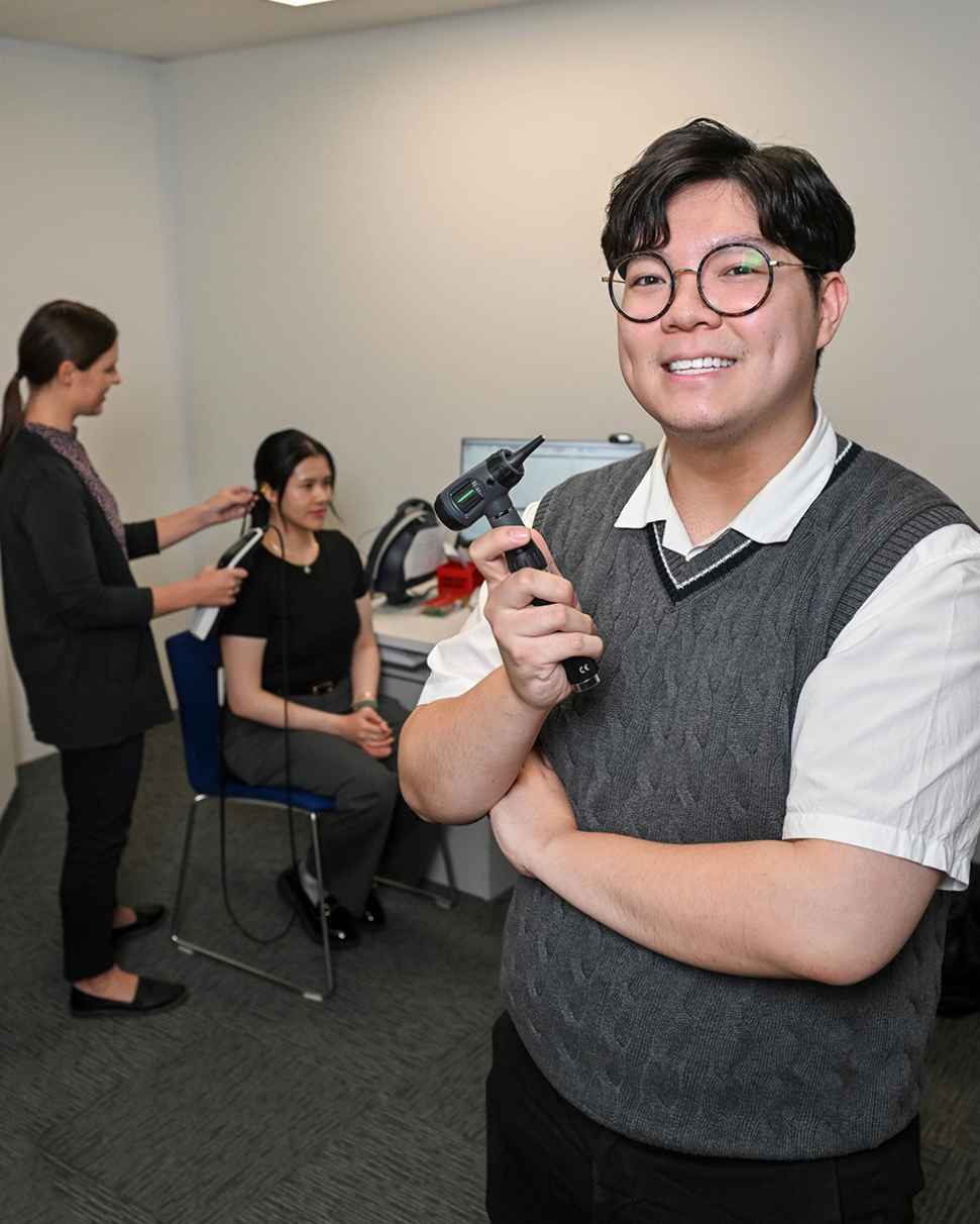 Flinders Audiology student stood smiling with tool with patient in the background