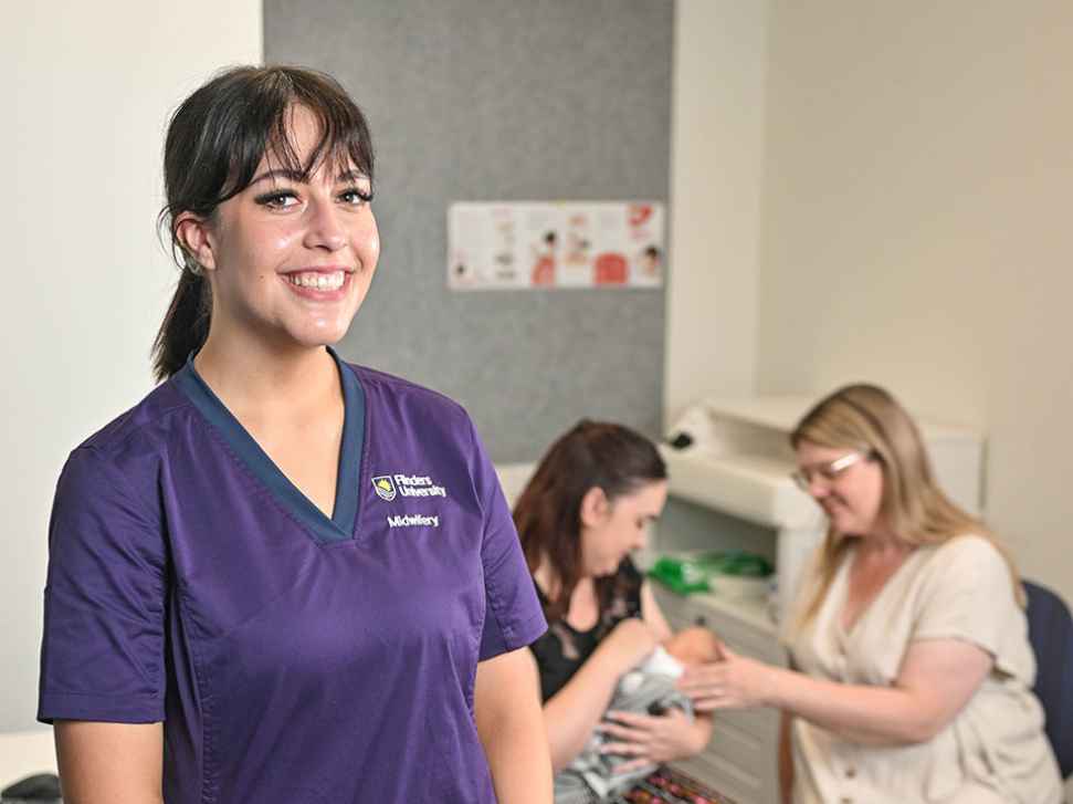 A Flinders Midwifery Student in a purple shirt stands beside a woman in a clinic, providing care and support.