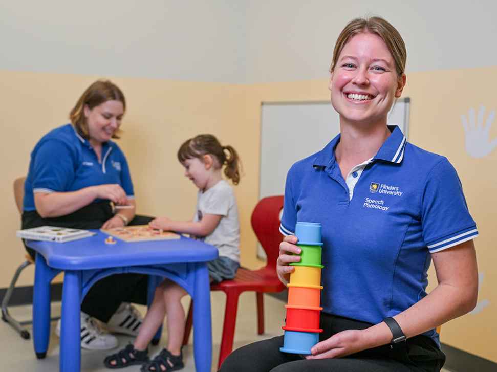 Flinders Speech Pathology Student smiling and holding colourful stacking toy with child in background