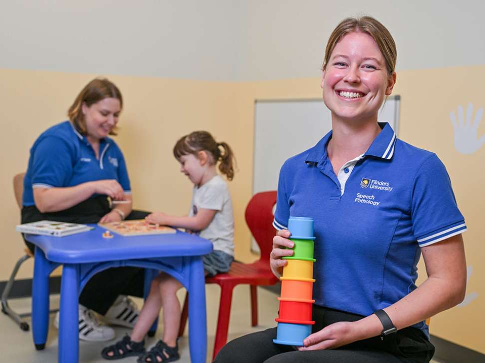 Flinders Speech Pathology Student smiling and holding colourful stacking toy with child in background
